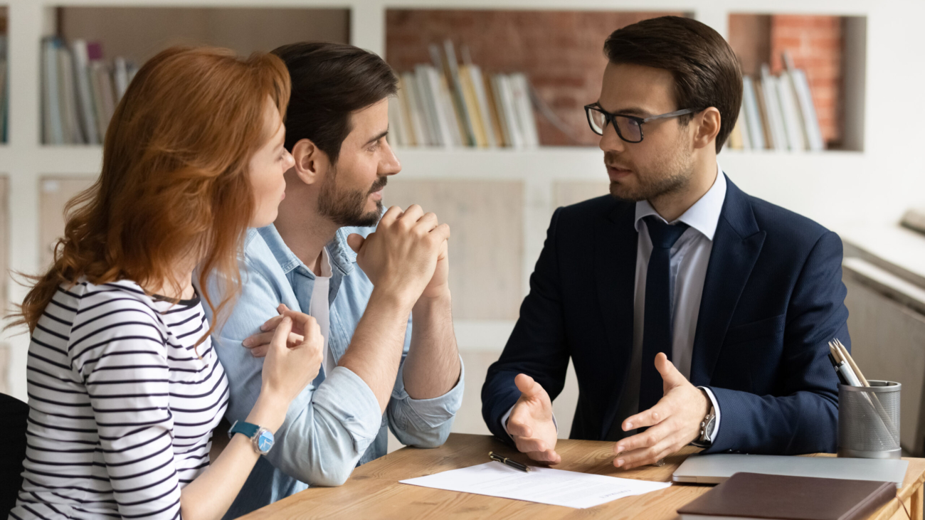 Professional male lawyer financial advisor consulting happy family couple clients in modern office. Interested young customers meeting Real Estate Agent broker bank worker, discussing agreement or deal.